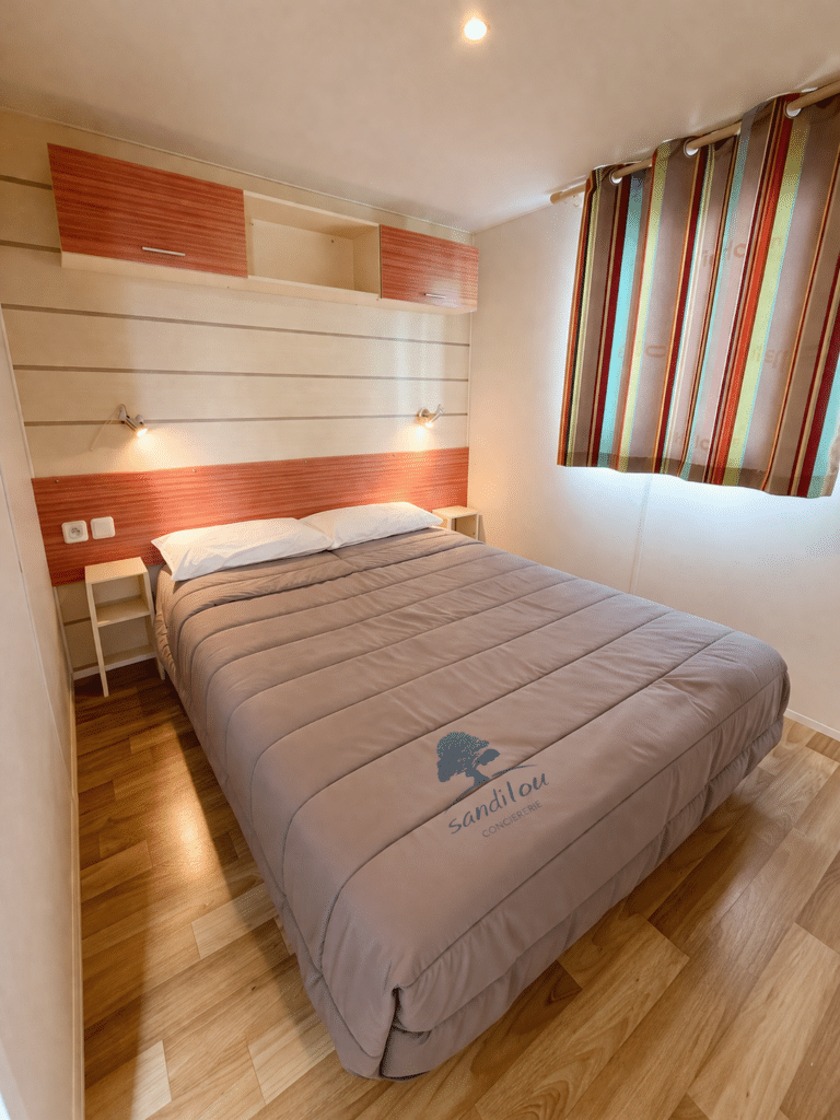Chambre de vacances avec lit double, couette grise Sandilou Conciergerie, rideaux colorés et mobilier en bois clair.
