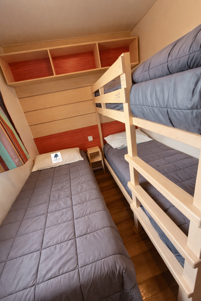 Chambre de mobil-home avec lit simple et lits superposés en bois, couettes grises et étagères oranges.