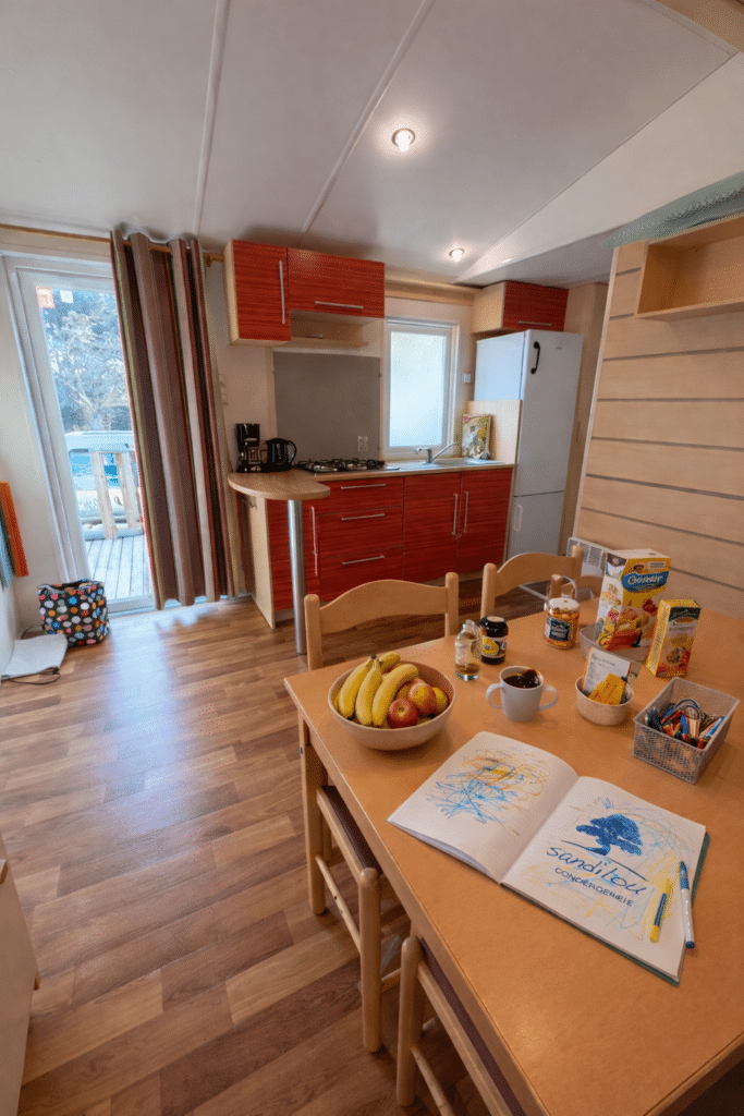 Intérieur chaleureux d'un mobil-home avec cuisine rouge et carnet sanditou CONCIERGERIE sur la table en bois.