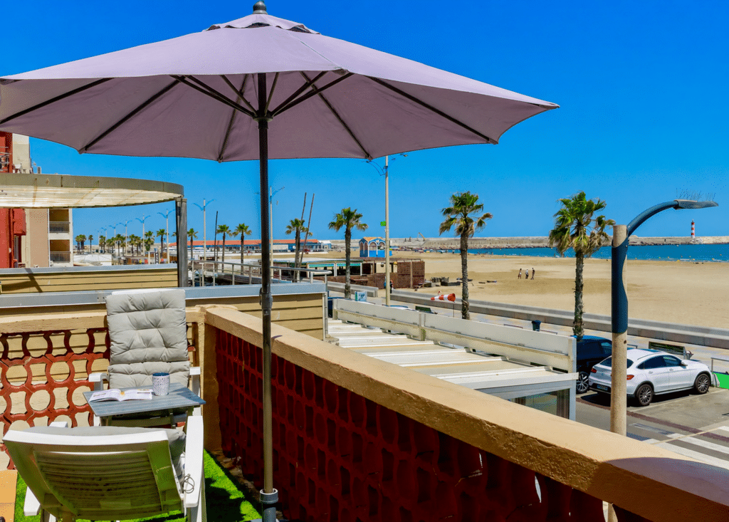 Balcon vue mer avec parasol violet et plage ensoleillée Balcon ensoleillé avec parasol violet vue sur mer, plage de sable, palmiers et ciel bleu azur.